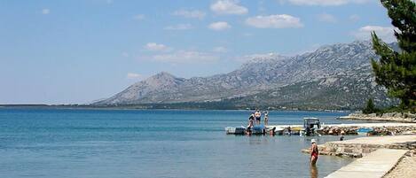 A coastal scene with a clear sky, calm sea, rocky shoreline, and a mountain in the background.