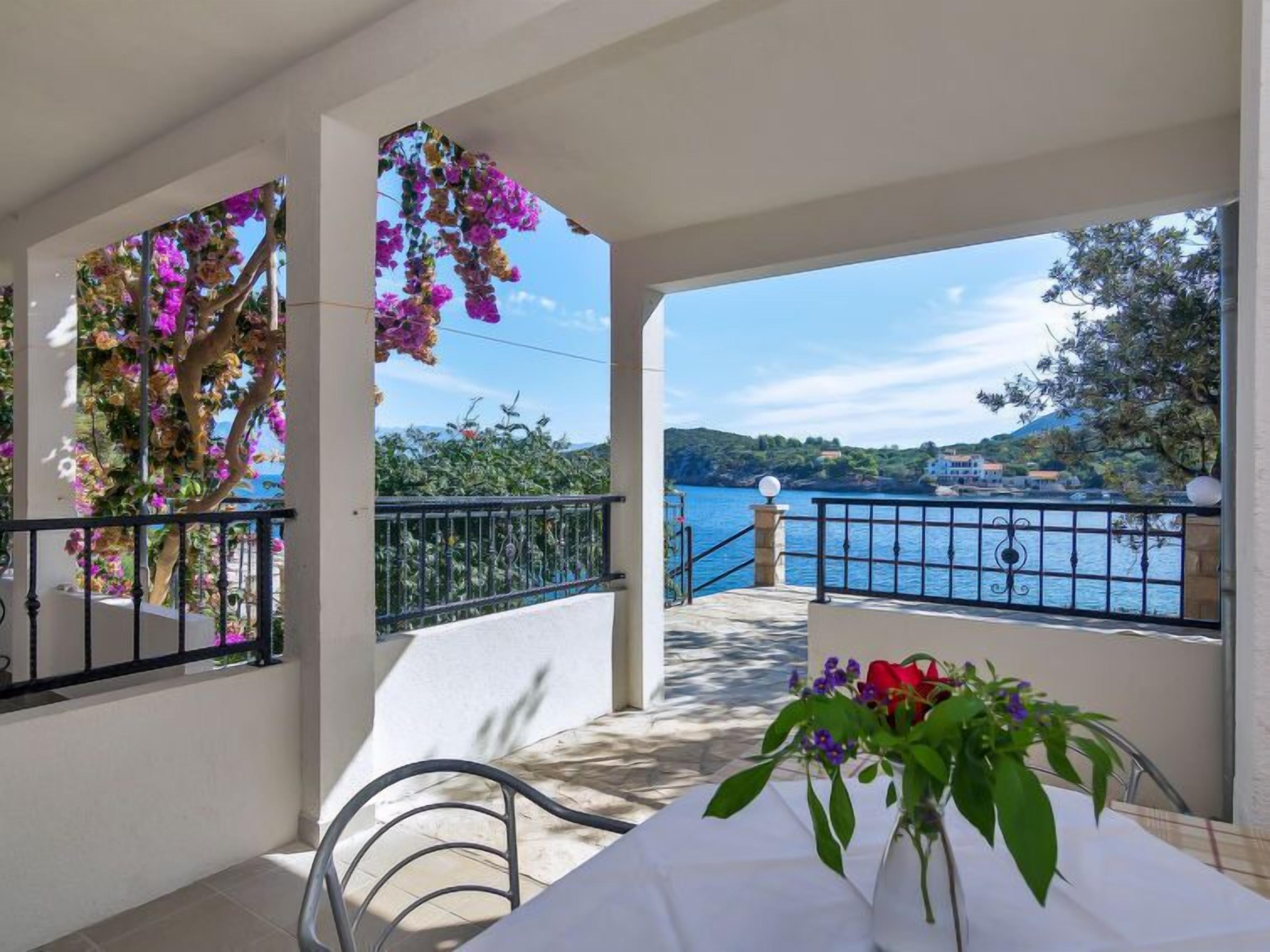 A balcony with a view of a lake and mountains, a table with a vase of flowers, and a chair.