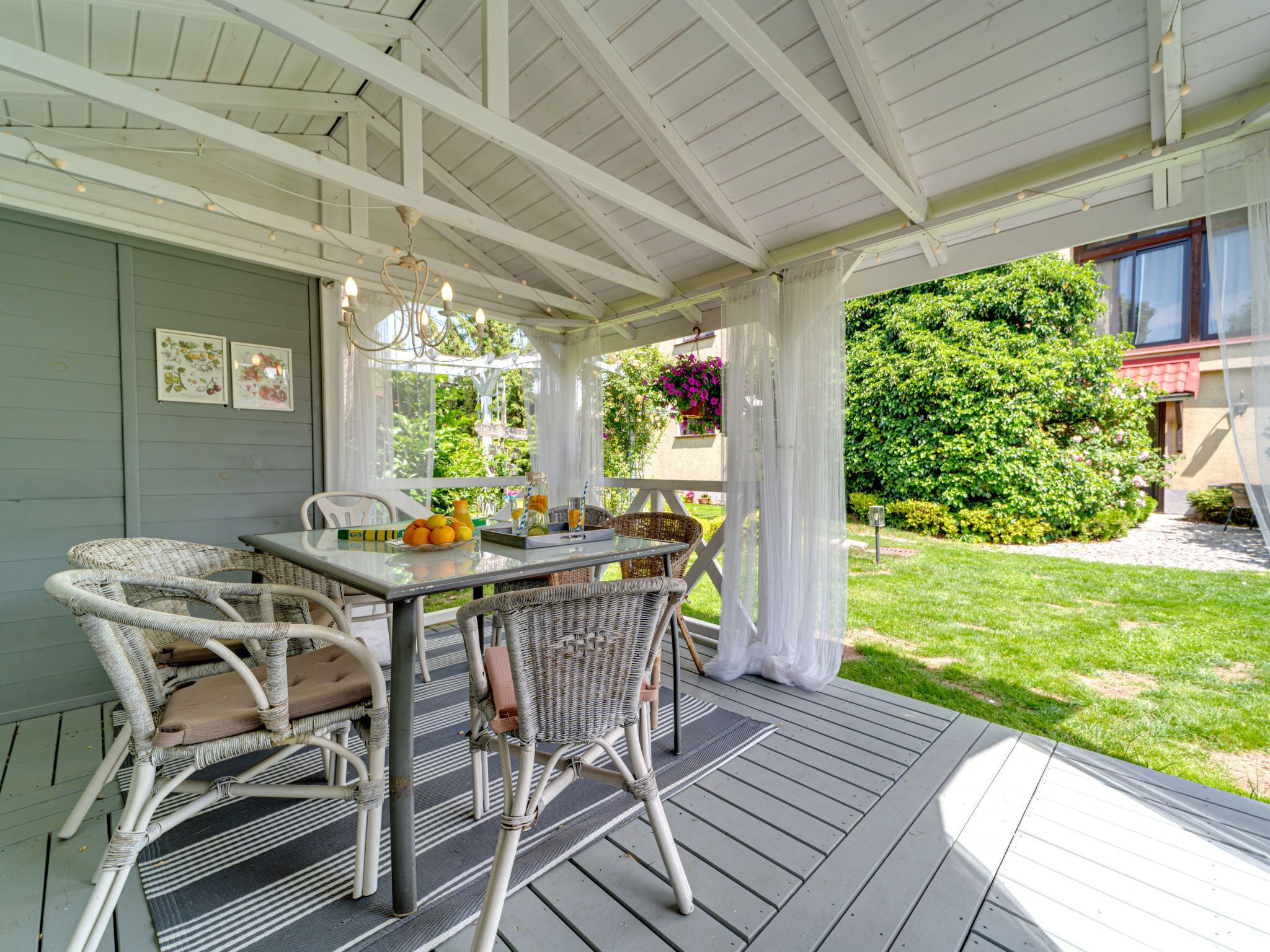 A covered outdoor patio with a table and wicker chairs, surrounded by greenery and a house in the background.