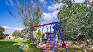 A playground with a swing set and climbing structure in a grassy area with trees and a house in the background. - Apartment B&B by Interhome (Medulin/Ližnjan)