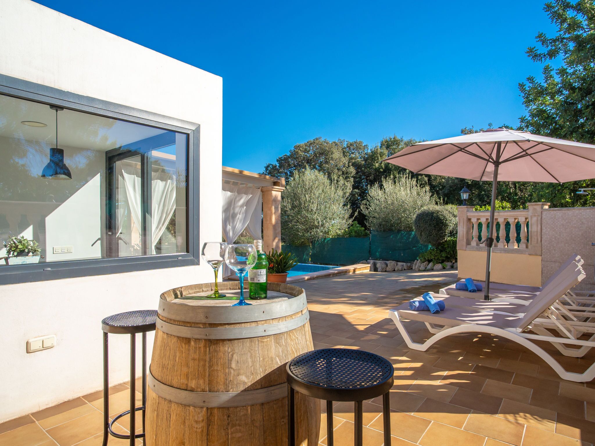 A patio with a wooden barrel, two stools, a lounge chair, and an umbrella.