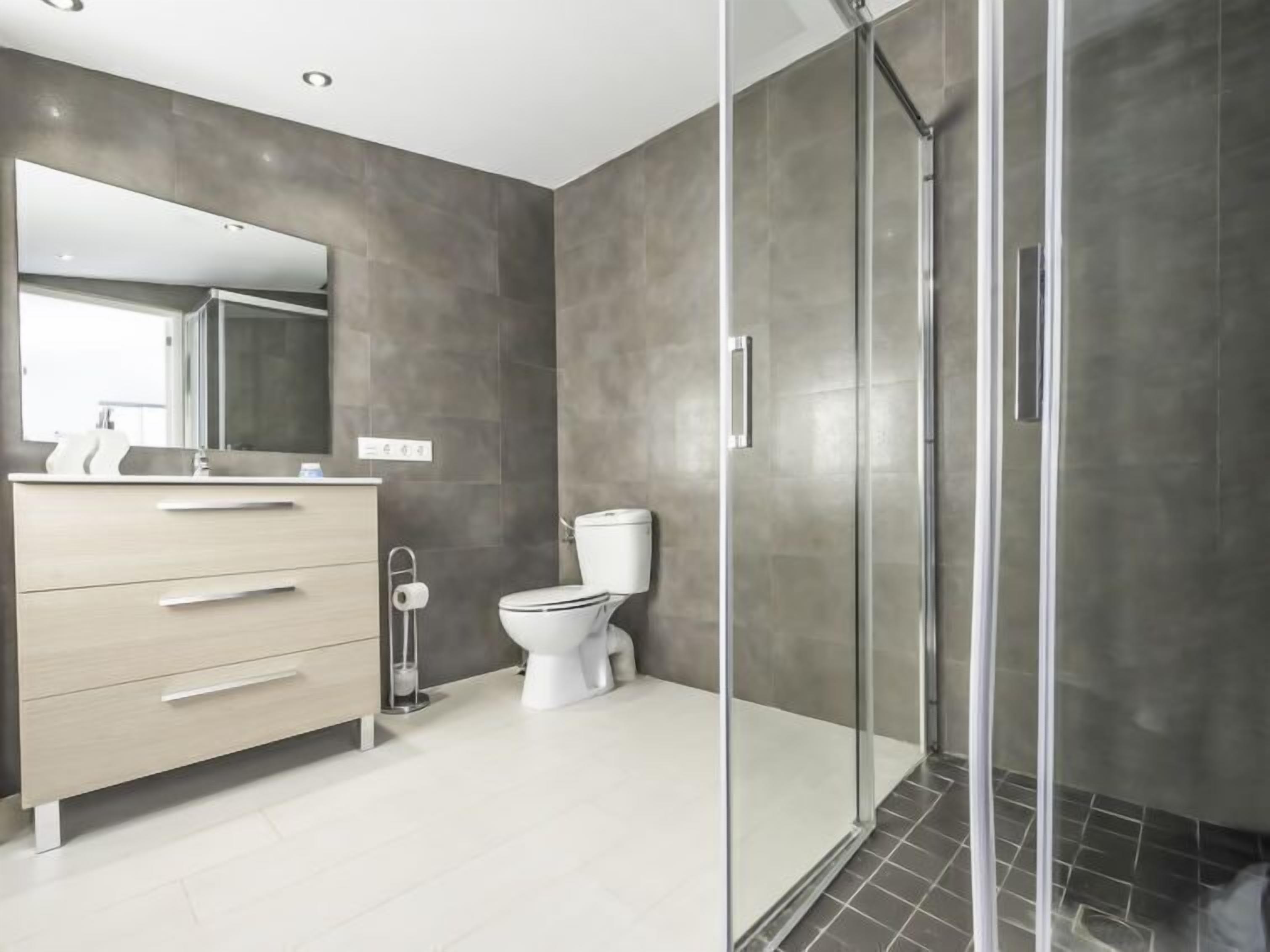 Une salle de bain moderne avec une douche à parois de verre, des toilettes blanches et un meuble-lavabo de couleur claire agrémenté d’un miroir.
