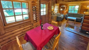Interno di una baita in legno con zona pranzo, soggiorno con divano e camino, e ampie finestre che offrono vista sull'esterno.