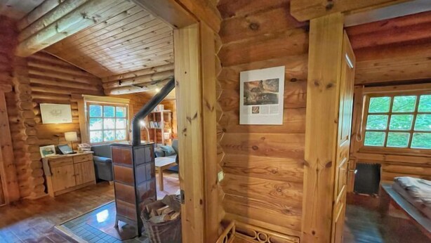 Accogliente interno di una baita in legno con camino, pareti di legno, una scala e una finestra che si affaccia sul verde.