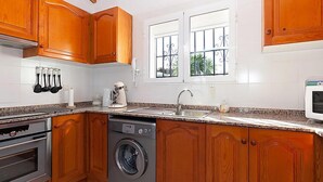 Eine Küche mit Holzschränken, einem eingebauten Backofen, einer Waschmaschine, einer Mikrowelle und einem Fenster mit Gitterstäben.