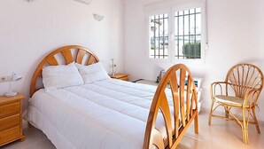 Ein Schlafzimmer mit einem hölzernen Bett, einer Nachttischlampe, einem Sessel und einem Fenster mit Gitterstäben.