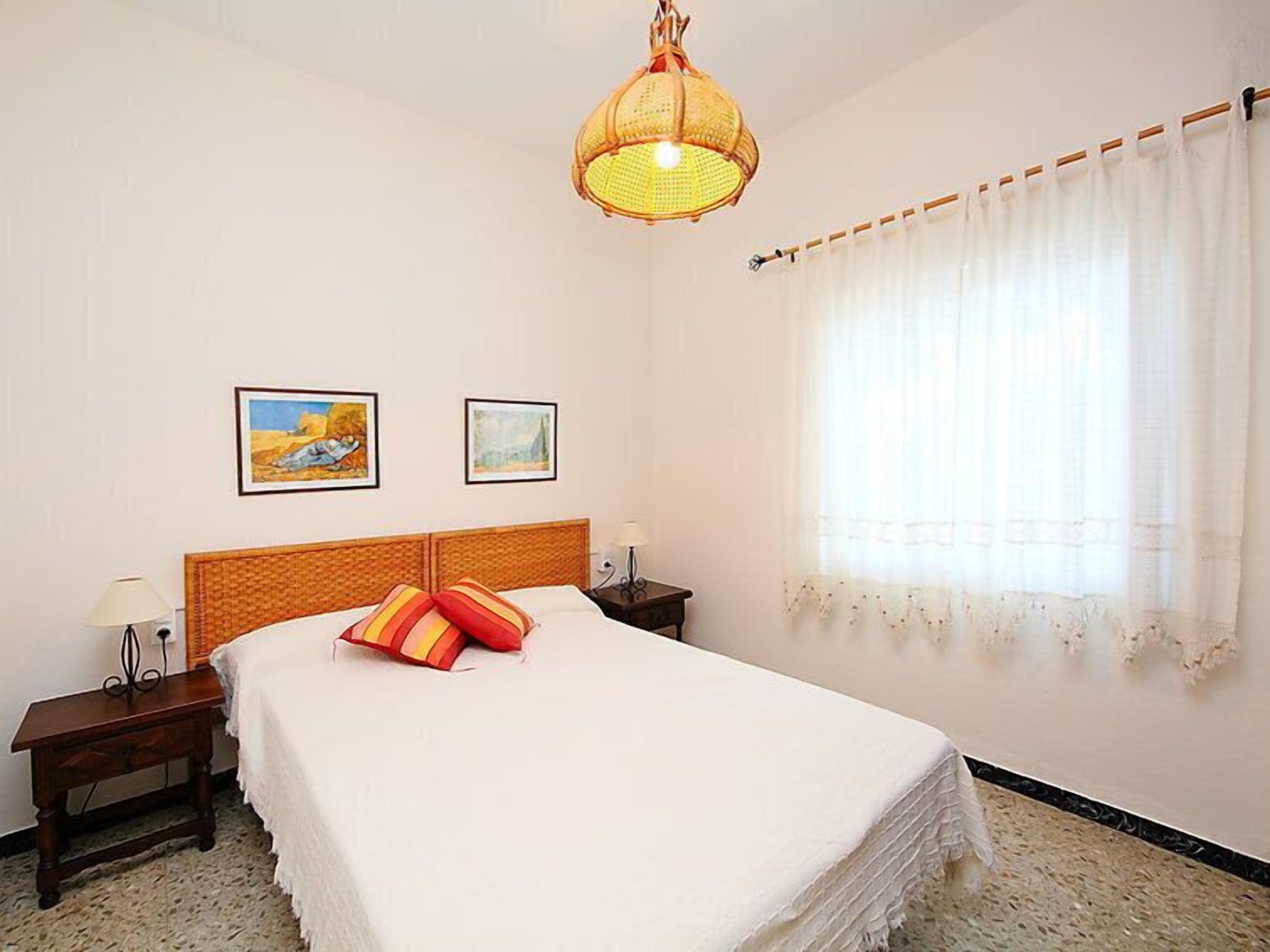 Habitación de hotel con cama, dos mesitas de noche, una lámpara colgante, ventana con cortinas y dos cuadros en la pared.