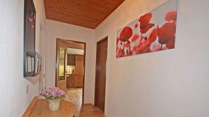 Un passadís amb sòl de fusta, una taula de fusta amb un ram de flors, una làmpada fixada a la paret i una porta que dona a una zona de cuina.