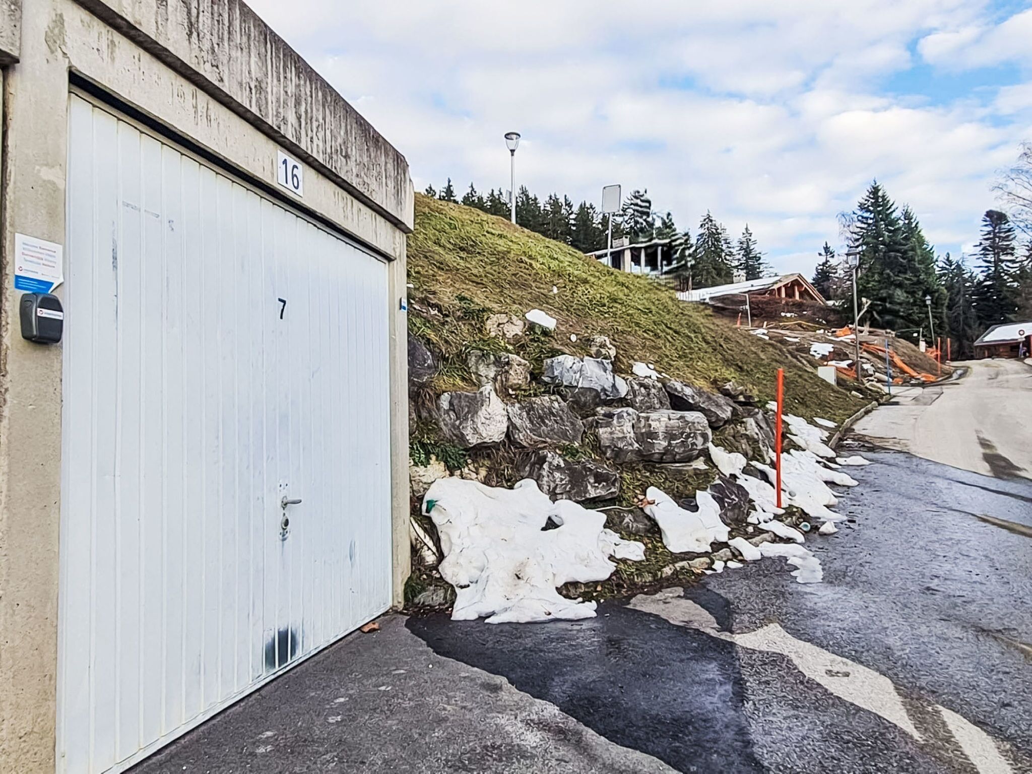 Ein Gebäude mit einer weißen Garagentür, die mit den Nummern 16 und 7 versehen ist, neben einer Steinmauer mit Schneeflecken.