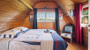A cozy cabin bedroom with a wooden ceiling, a bed with a nautical-themed bedspread, a window with curtains, and a small table with a chair.