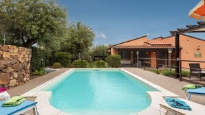 A rectangular swimming pool with a stone wall on one side and a covered patio area with lounge chairs.