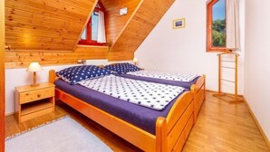 Ein Schlafzimmer mit schräg laufender Decke, Holzwänden, zwei Betten mit sternenmustiger Bettwäsche, einem Nachttisch, einer Lampe, einem Fenster mit Vorhängen und einer Holzleiter.