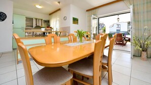 Een eetgedeelte met een houten tafel en stoelen, een keuken op de achtergrond en uitzicht op het groen door een glazen deur.