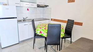 Kompakti keittiö, jossa on valkoiset kaapit, sisäänrakennettu uuni ja pesuallas. Keittiössä on ruokapöytä, jonka päällä on värikäs pöytäliina, sekä kaksi tuolia.