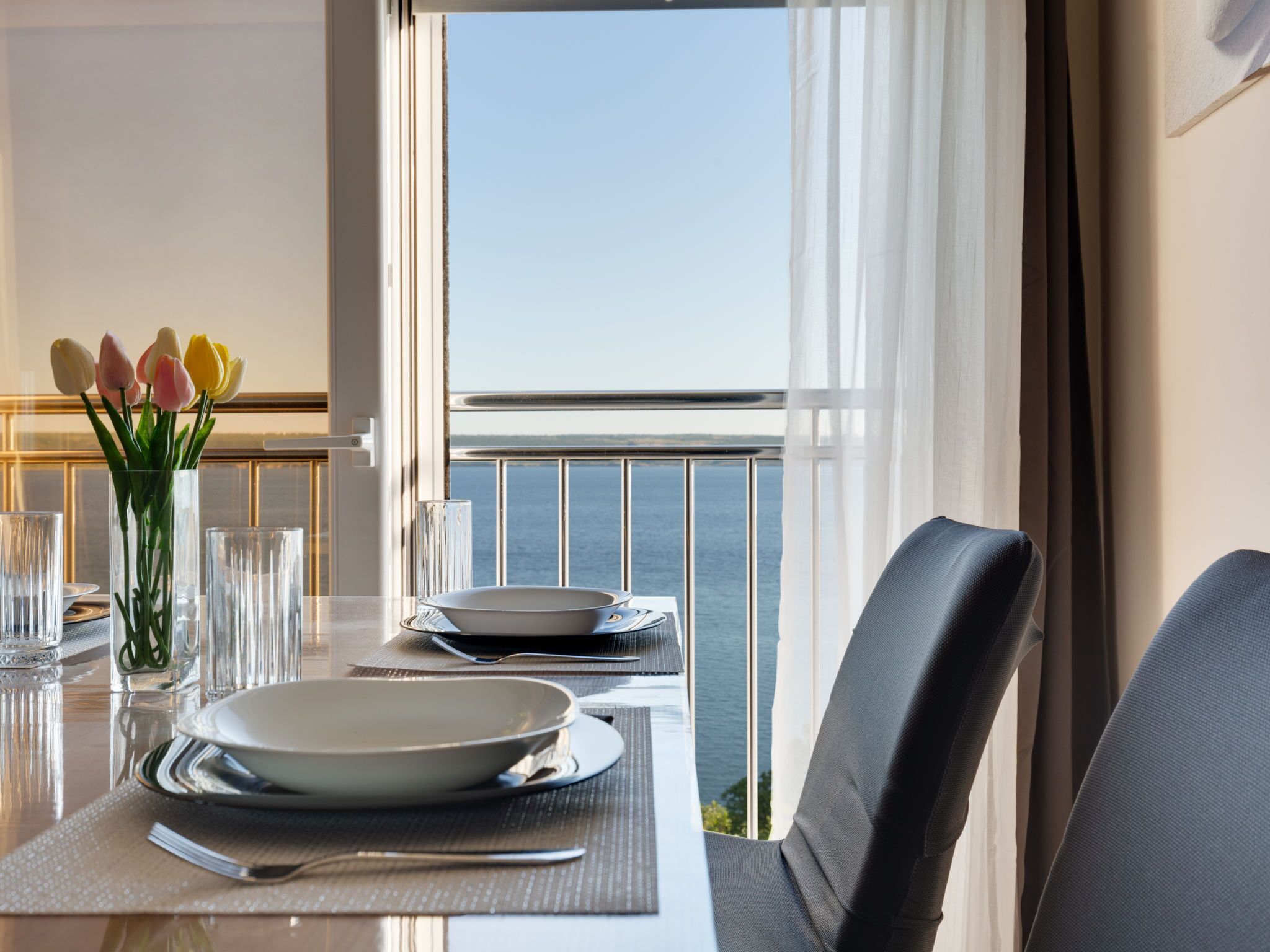 A dining table with plates, cutlery, and a vase of tulips, set against a balcony with a view of the sea.