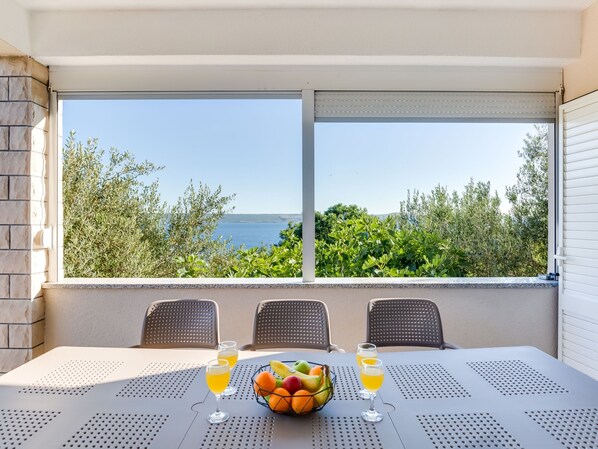 A table with two chairs, a fruit bowl, and two glasses of orange juice, overlooking a scenic view of the sea and greenery.