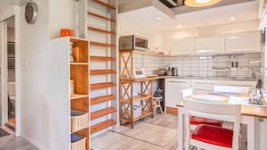 A modern kitchen with white cabinets, a wooden shelf unit, and a dining area with a red chair. - Apartment Rekowo by Interhome (Rekowo)