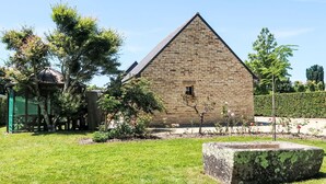 Un bâtiment en pierre doté d’un toit triangulaire, entouré d’un jardin bien entretenu où poussent diverses plantes, et agrémenté d’un grand élément décoratif en pierre au premier plan.