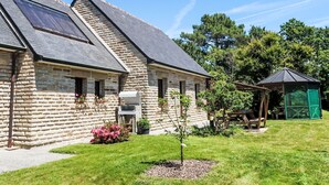 Un bâtiment en pierre avec un toit en pente et des panneaux solaires, un petit jardin avec un arbre et des fleurs roses, et un kiosque en arrière-plan.