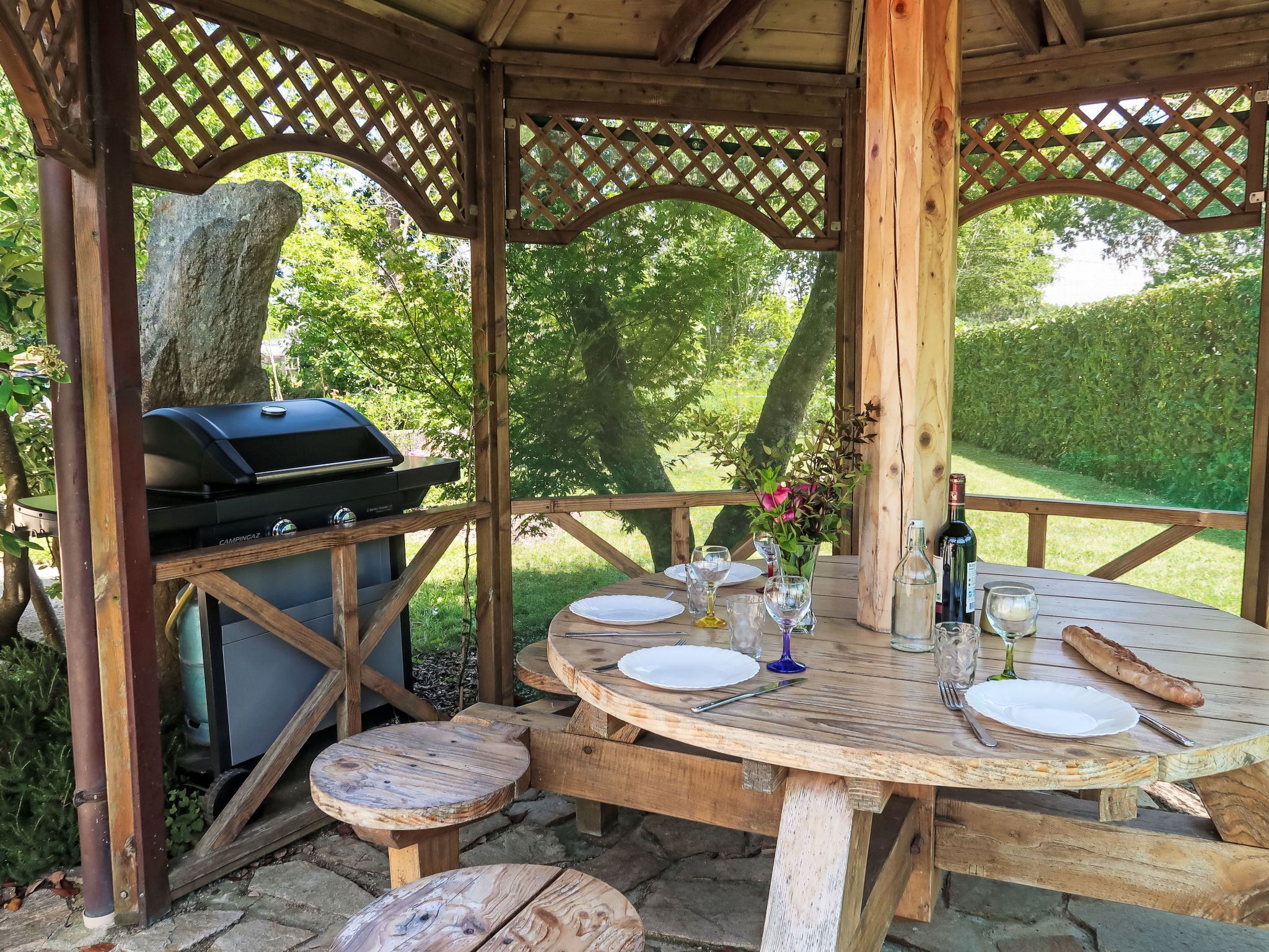 An outdoor wooden gazebo with a grill, a round wooden table set for four, and a vase with flowers.