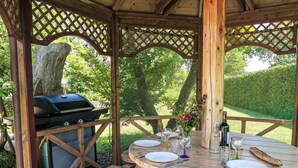 Un belvédère en bois installé à l’extérieur, doté d’un barbecue, d’une table ronde en bois pouvant accueillir quatre personnes et d’un vase rempli de fleurs.