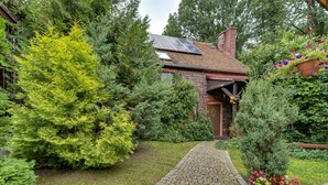 Une maison en briques avec un chemin pavé menant à l’entrée, entourée d’une végétation luxuriante et agrémentée d’un pot de fleurs suspendu.