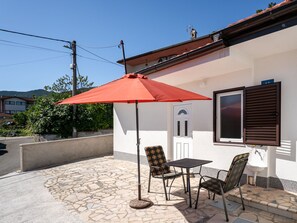 Een terras buiten met een tafel en stoelen, een rode parasol en een wit gebouw met een raam en een deur.