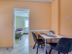 A room with a dining table, two chairs, a bed with a purple pillow, a nightstand, and a window.