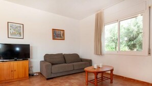 Un salon avec un canapé, un téléviseur posé sur un meuble en bois, une petite table ronde avec deux tasses, et une fenêtre avec des rideaux.