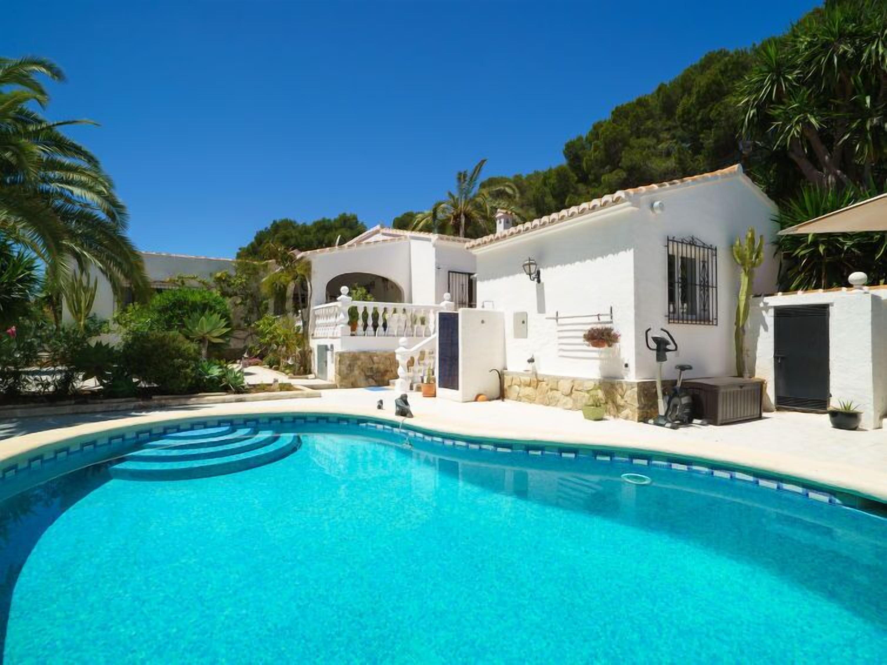 Vista de una piscina frente a una villa blanca con fachada de piedra, una pequeña escalera que conduce a un balcón y un exuberante jardín con palmeras.