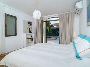 A bedroom with a bed, a bedside table, a lamp, a window, a bookshelf, and an air conditioning unit.