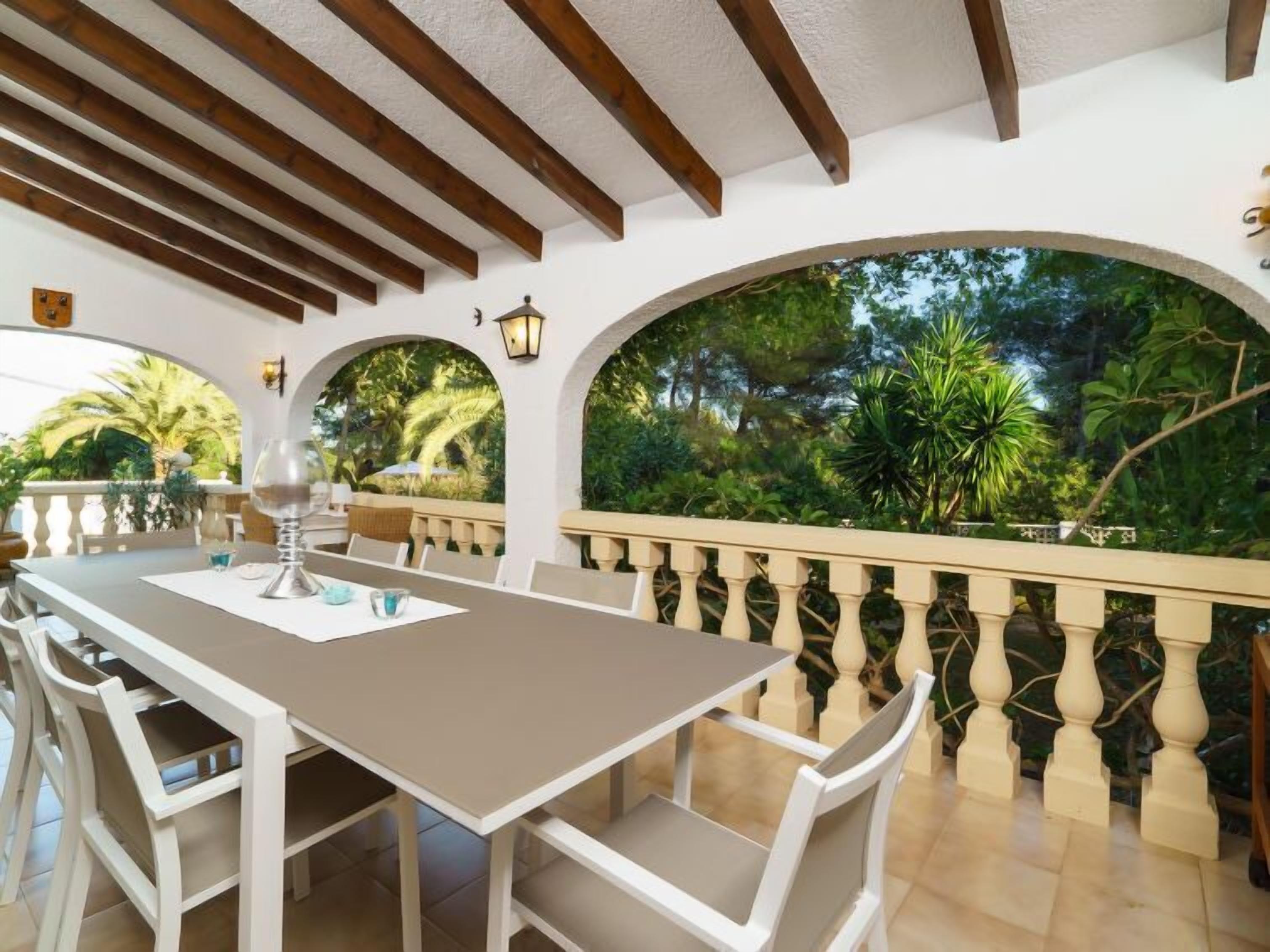 An outdoor dining area with a table and chairs, surrounded by a balustrade and a view of greenery.