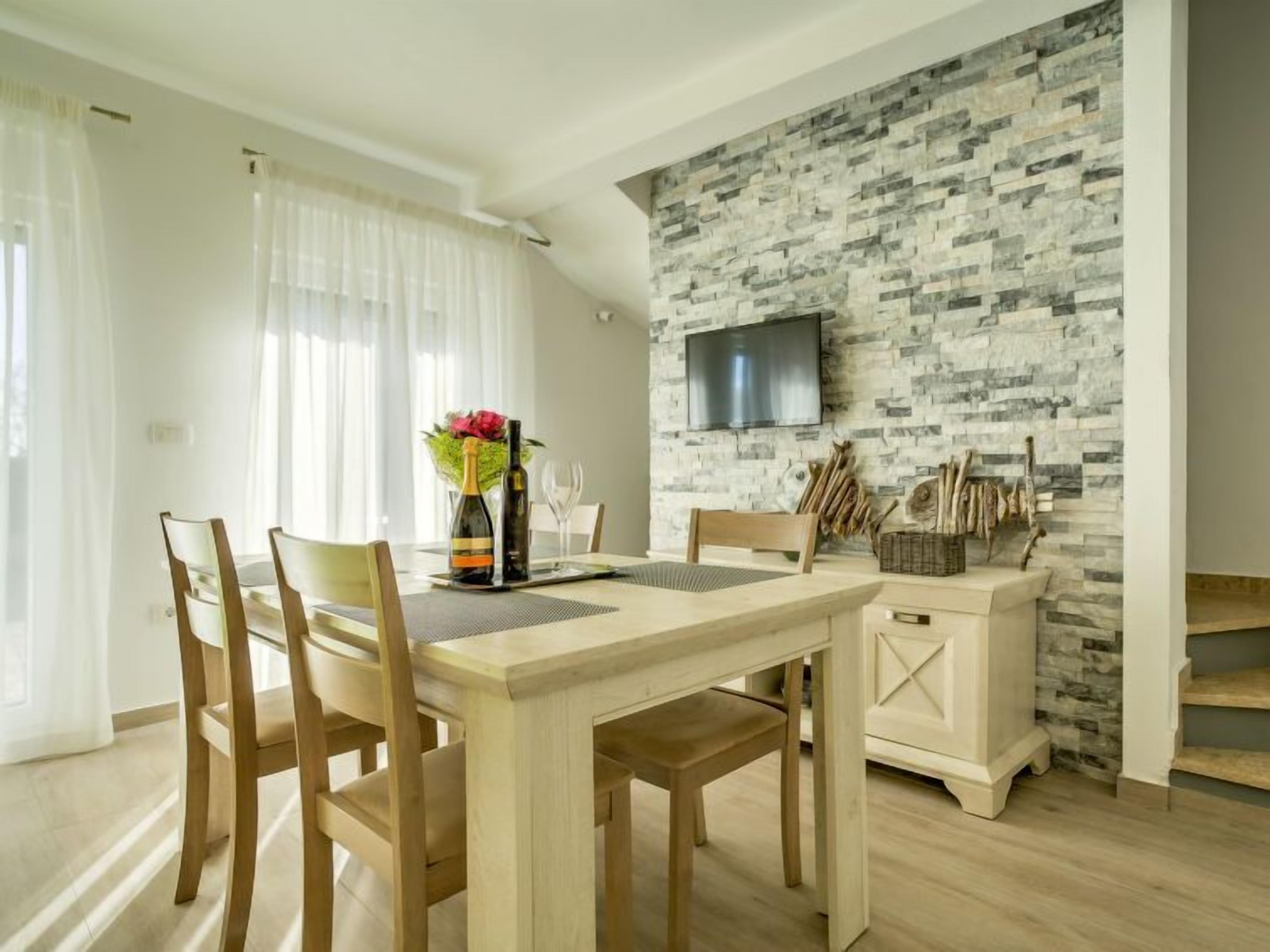 Un coin repas avec une table en bois, des chaises, un vase de fleurs et un téléviseur fixé à un mur en pierre.