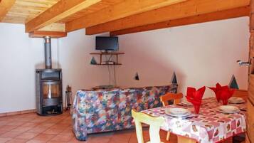 A dining area with a wooden table, chairs, and a tablecloth. There is a fireplace and a television on a shelf.