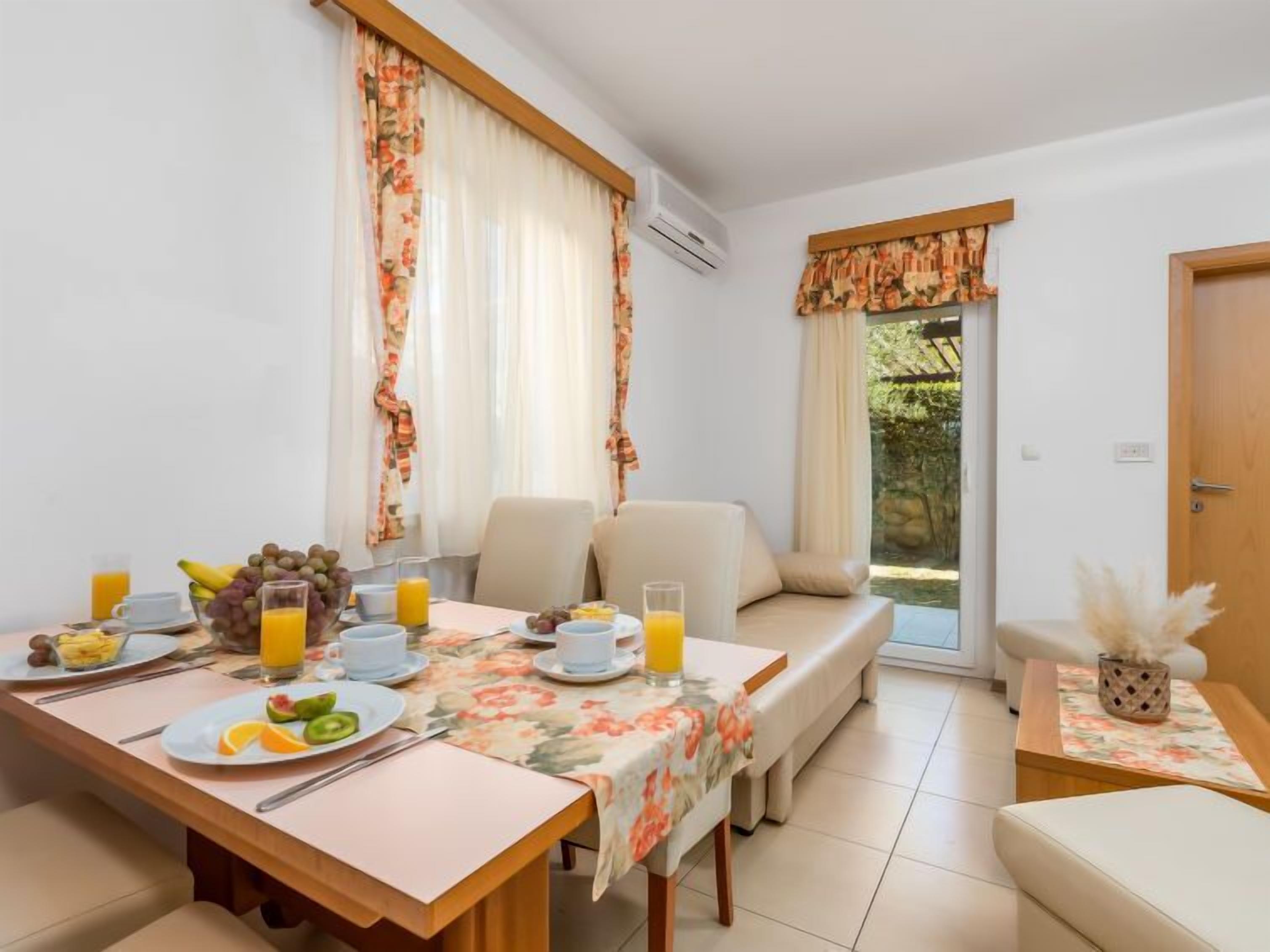 Ein Essbereich mit einem für das Frühstück gedeckten Tisch, mit Früchten und Saft, sowie eine Couch mit Blick auf Grünflächen.