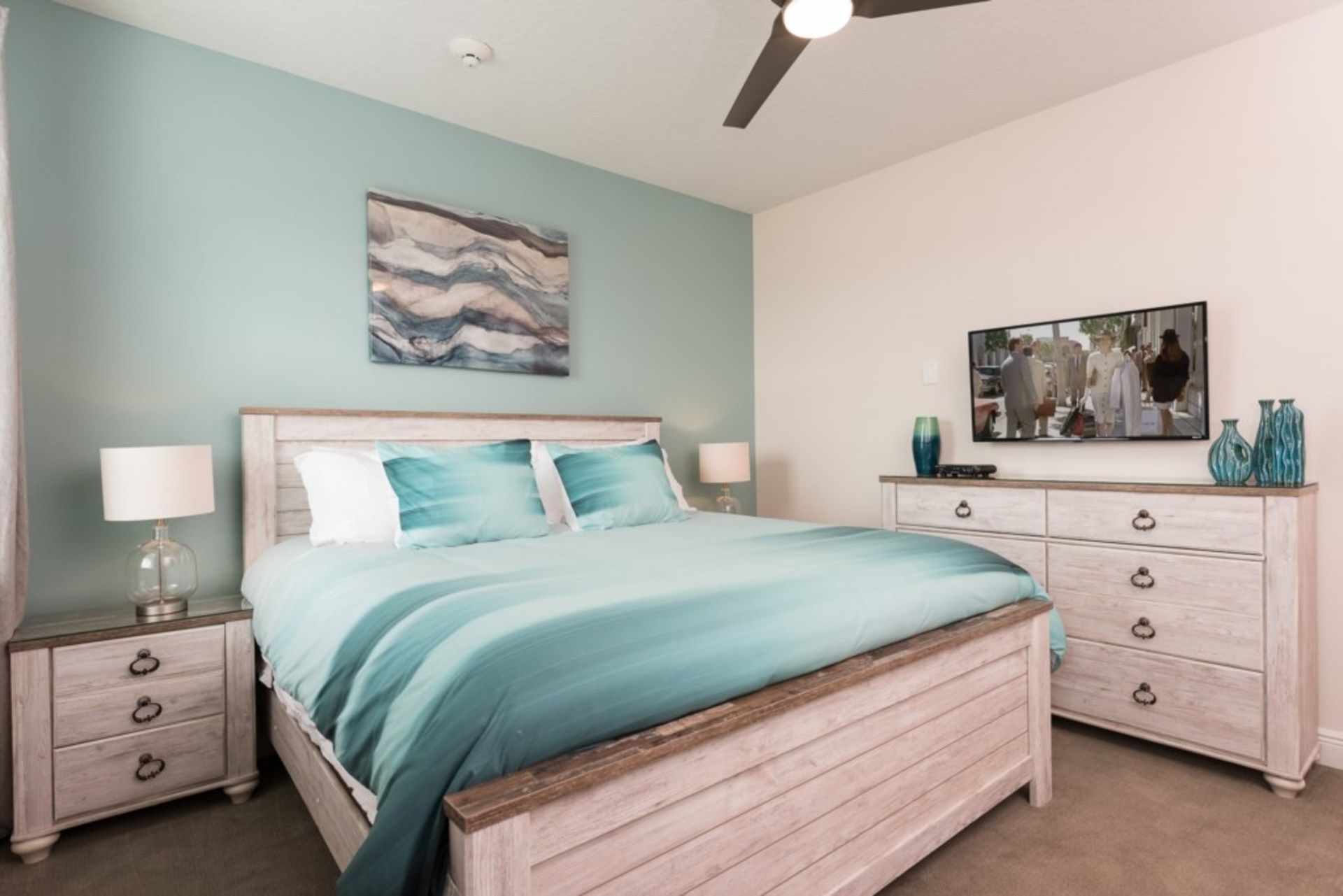 A bedroom with a bed, nightstands, a dresser, a ceiling fan, and artwork on the walls.