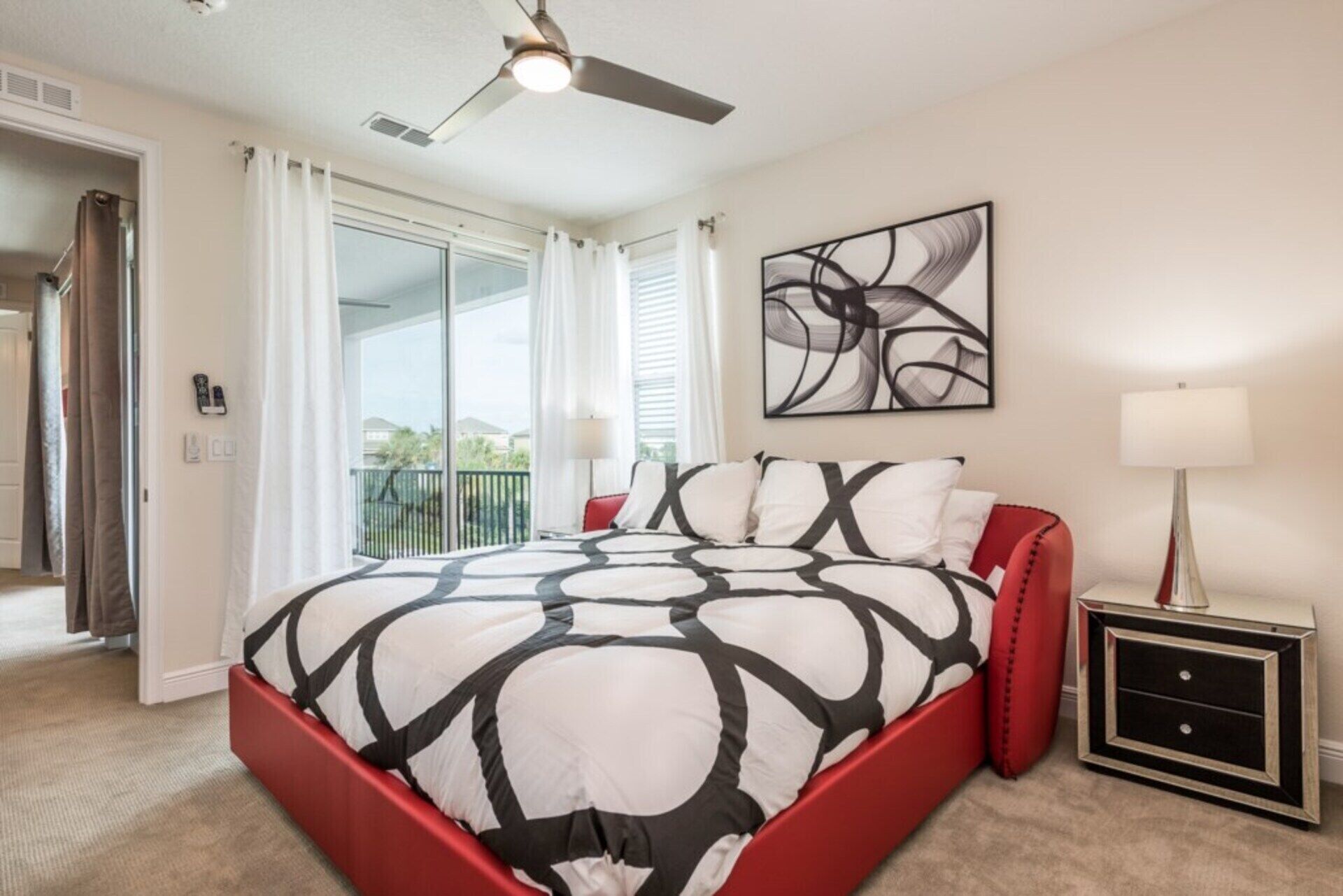 A bedroom with a bed, a ceiling fan, a painting, a nightstand, and a view of the outside.