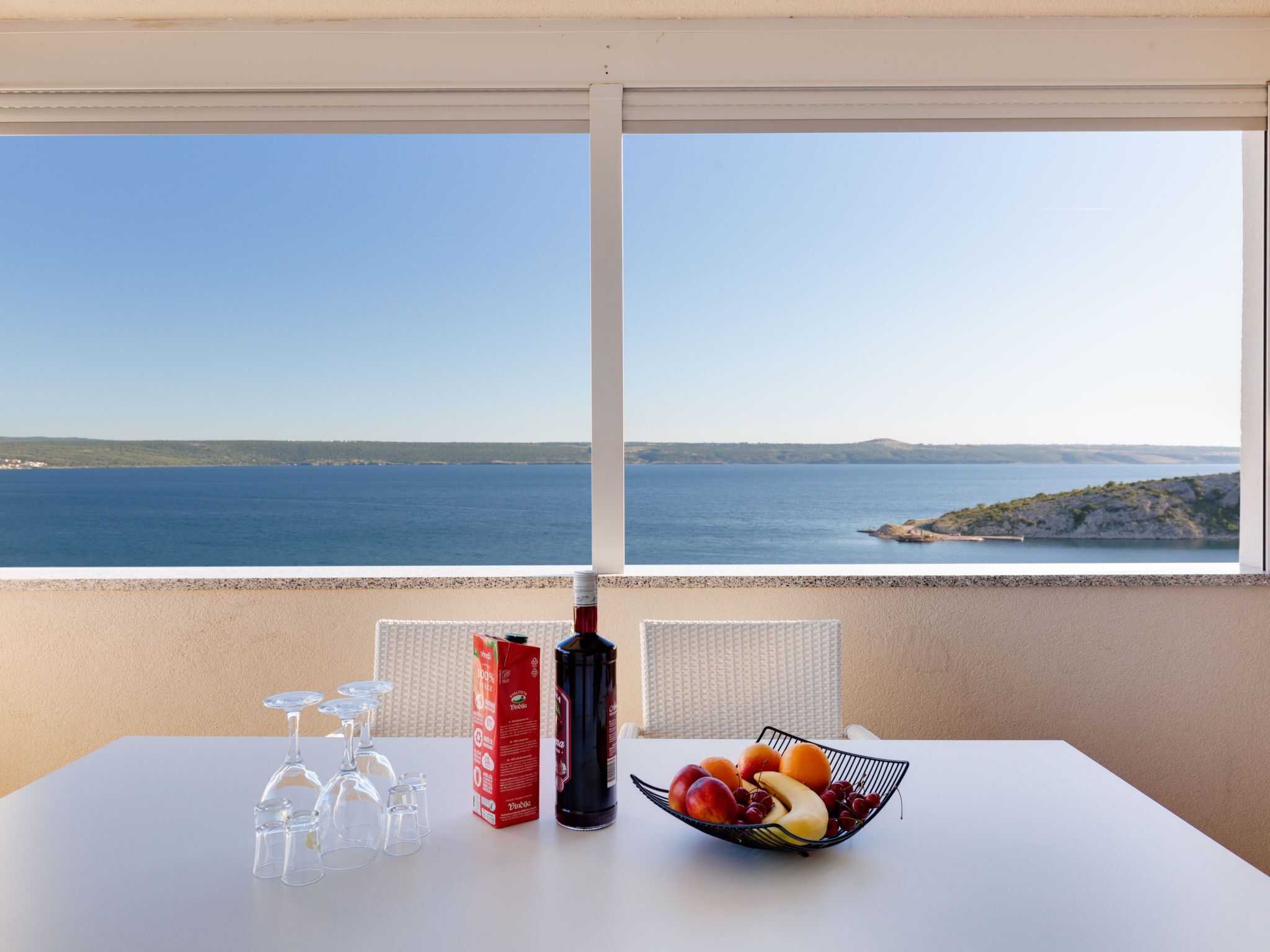 A table with a fruit bowl, wine glasses, and a bottle of wine, overlooking a coastal view.