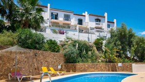 Una piscina con sillones de descanso, un muro de piedra y un edificio con balcones al fondo.