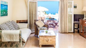 Habitación de hotel con cama, sofá, mesa de centro y televisor. Tiene un balcón con mesa y sillas, y vista al mar.