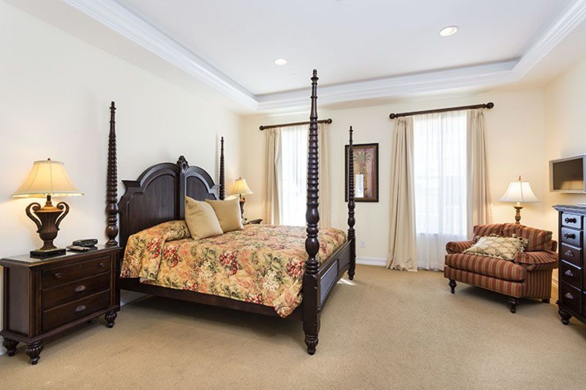 A four-poster bed with floral bedding, a wooden nightstand with a lamp, a brown leather sofa, and a television on a dresser.
