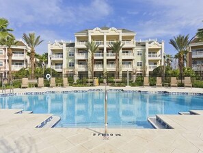 A swimming pool with a clear blue water surface, surrounded by a tiled deck and a fence. In the background, there is a multi-story building with balconies and palm trees. - Private Villa 1279 (Kissimmee)