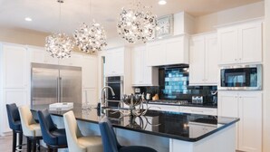 Cocina moderna con armarios blancos, una isla central con taburetes, electrodomésticos de acero inoxidable y elegantes candelabros de cristal.