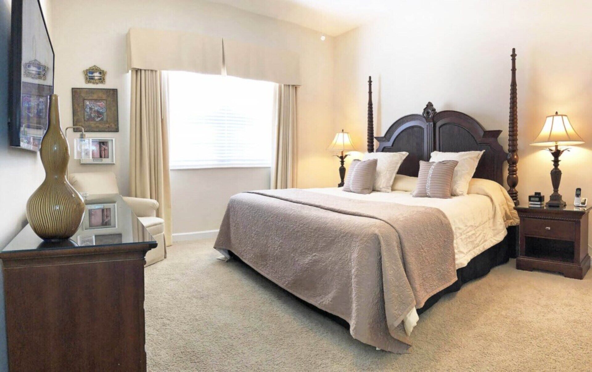 A neatly arranged bedroom with a large bed, two bedside lamps, a nightstand, a dresser, and a window with curtains.