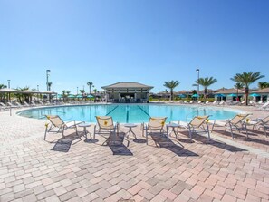 A swimming pool with lounge chairs and umbrellas, surrounded by palm trees and a covered area. - Villa in Champions Gate 1134 (Davenport)