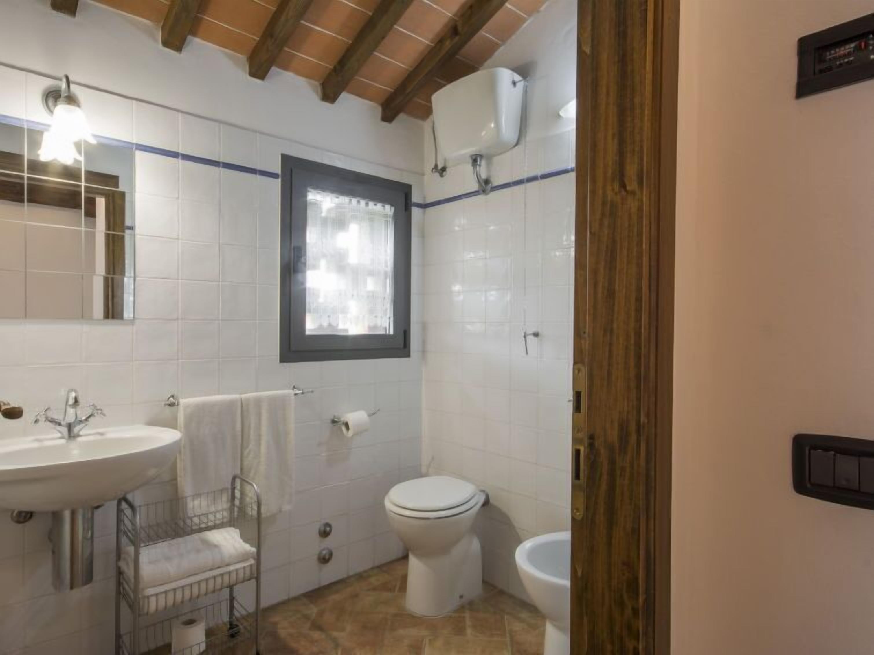A bathroom with a white sink, a toilet, and a bidet. There is a window with a curtain, and the floor is tiled.