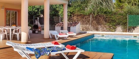 Un área de piscina con sillones reclinables y una mesa, rodeada por un porche cubierto y vistas a árboles.