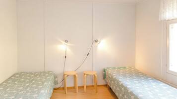 Une chambre avec deux lits simples, un petit tabouret en bois, un tapis tissé et un luminaire fixé au mur.