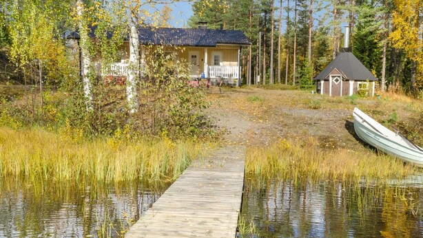 A wooden dock leading to a house surrounded by tall trees and a small boat docked nearby.