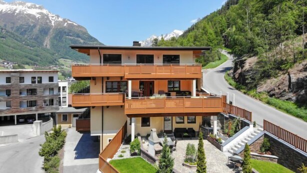 Un immeuble à plusieurs étages doté d’un balcon, entouré de verdure et avec des montagnes en arrière-plan.